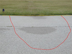 Photo showing a large-severity depression on the edge of an asphalt pavement. A stringline has been stretched across the depression to illustrate the depth, and some water staining on the pavement surface is noticeable at the center of the depression. A red line has also been drawn on the photo to outline the area with the depression.