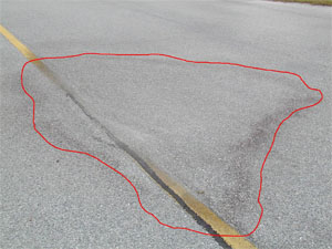 Photo showing a medium-severity depression on the pavement. The area of the depression is noticeable due to water staining on the pavement. A red line has also been drawn on the photo to outline the area with the depression.