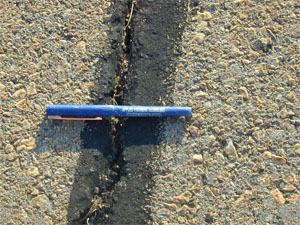 Close-up photo of a single medium-severity crack. The crack is unsealed, and appears to be slightly larger than 1/4 in (6 mm) wide. A ball point pen has been placed across the crack to give it scale.