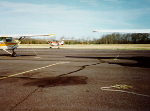 Overview photo of an apron with multiple larger areas of oil staining on the pavement surface. The photo also shows three smaller airplanes sitting on this apron near the stains.