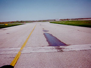 Overview photo of a taxiway pavement with visible low-severity rutting in one wheel path. The rutting is visible because there is standing water in the wheel path.