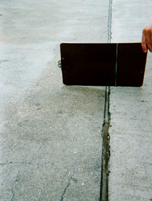 Close-up photo showing a transverse joint with a slight difference in elevation across the joint. A clipboard is placed across the joint in the photo to illustrate that there is a height differential between the two slabs.