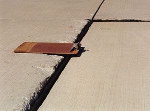 Close-up photo showing a transverse joint with a 1/2 in (13 mm) difference in elevation across the joint. A clipboard is placed across the joint in the photo to illustrate that there is a height differential between the two slabs.