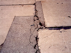 Close-up photo of an irregularly shaped asphalt patch within a PCC slab. The patch contains considerable visible distress that includes spalling, cracking, and a noticeable loss of material.